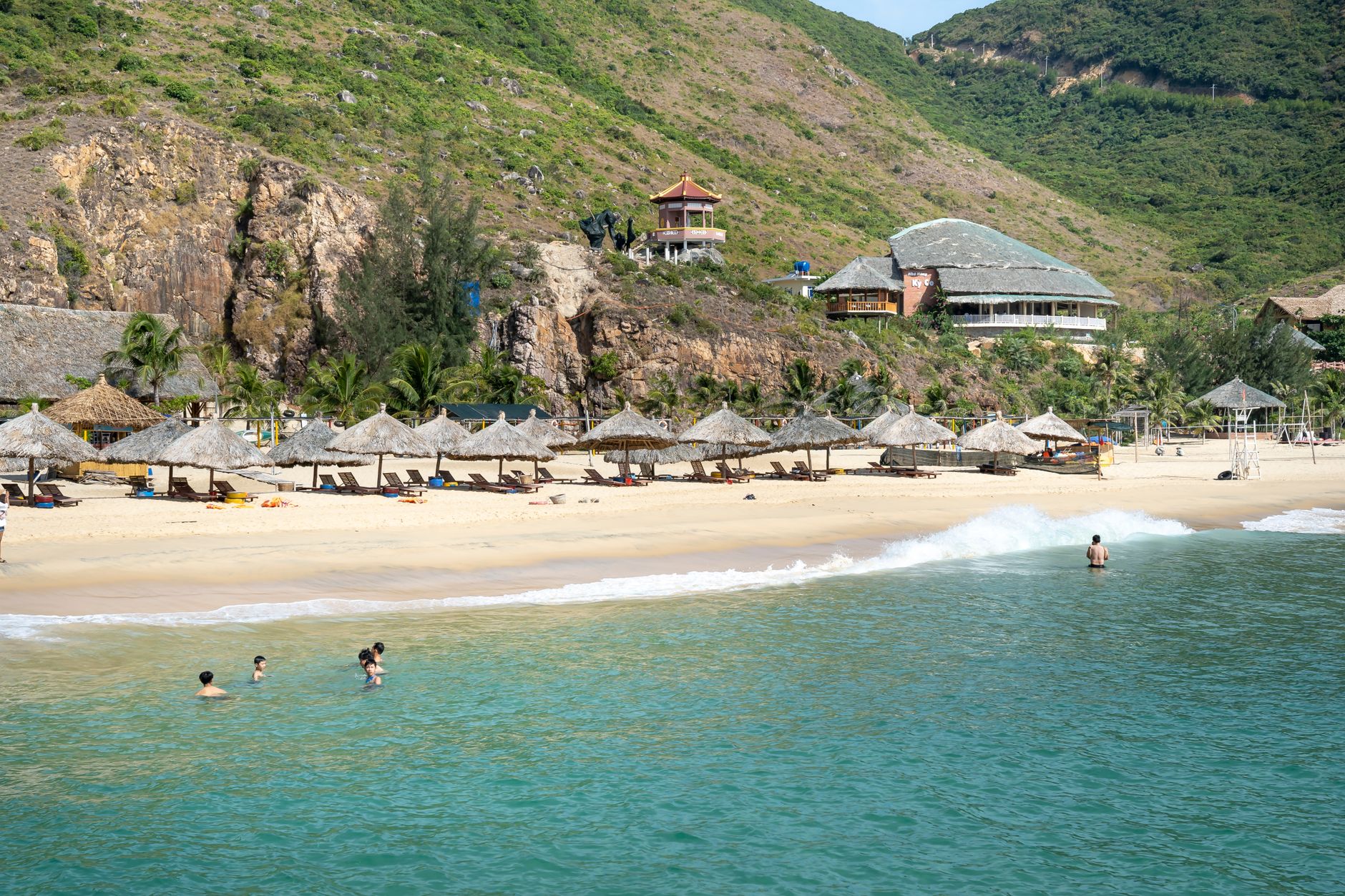 People swimming and relaxing on sandy beach with straw umbrellas surrounded by grassy rough hills on sunny summer day