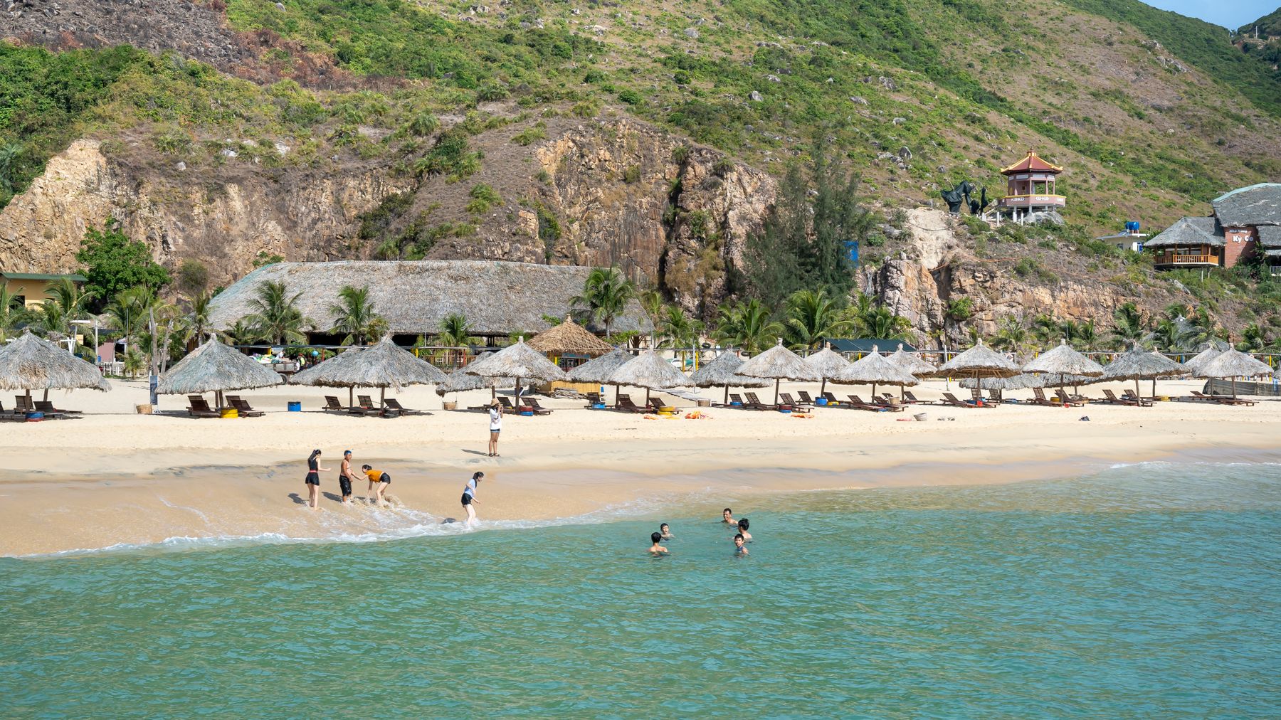 Menschen entspannen an einem Sandstrand mit Sonnenschirmen und Liegestühlen, umspült von türkisfarbenem Meerwasser und umgeben von struppigem Gras