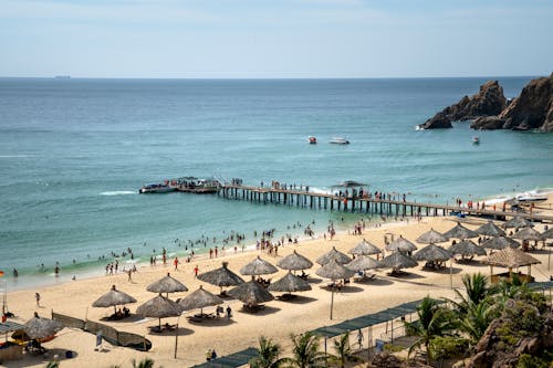 People chilling on spacious sandy beach with straw umbrellas and narrow wharf near turquoise rippling sea on sunny day