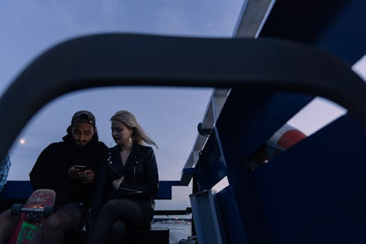 A young couple enjoying a tranquil boat ride under the evening sky, engrossed in a smartphone moment.