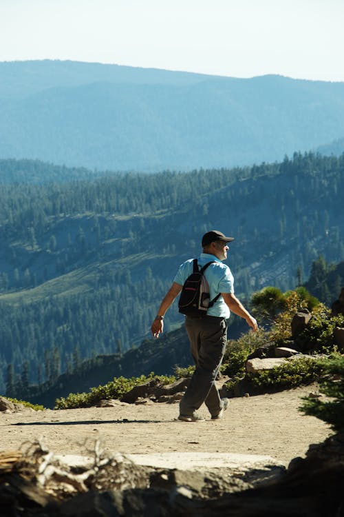 Free Side view of unrecognizable male traveler in casual clothes and backpack walking on trail during hiking in mountains covered with coniferous forests Stock Photo