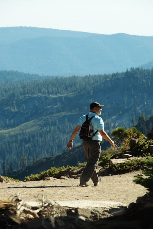 Side view of unrecognizable male traveler in casual clothes and backpack walking on trail during hiking in mountains covered with coniferous forests