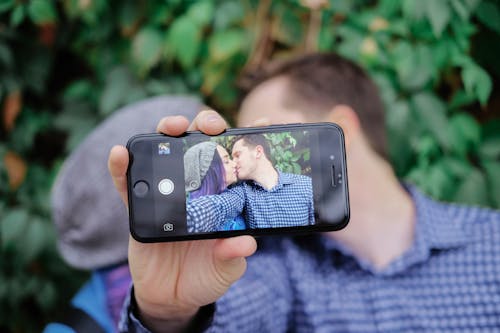 Romantic couple kissing tenderly while showing mobile phone screen with selfie while standing against lush park trees