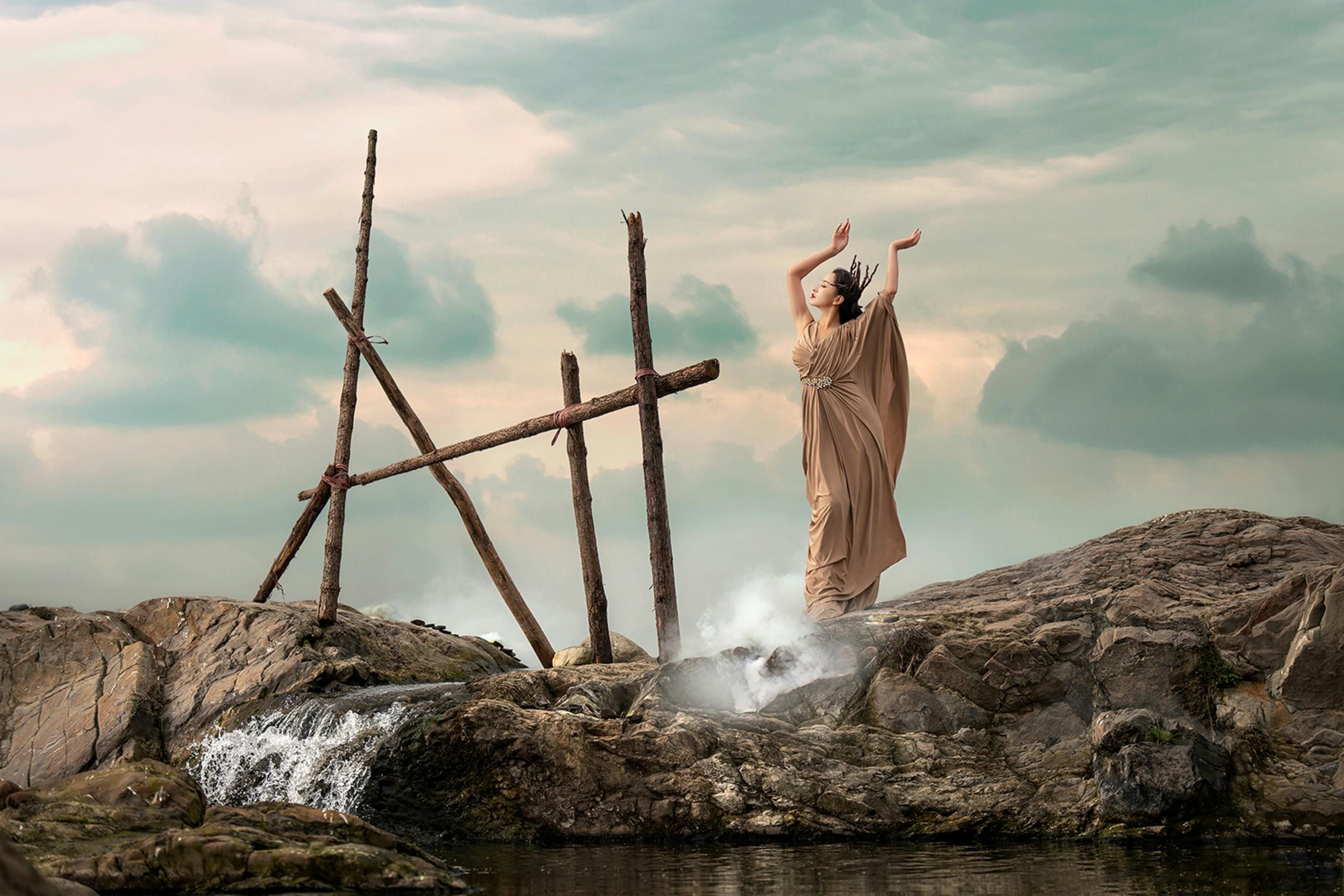 Full body of woman in long dress and crown standing on stone near rippled water and showing myth hero