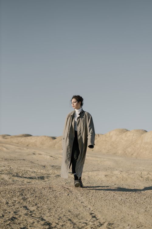 Free Elegant woman in trench coat strolling through a serene sandy desert landscape under clear skies. Stock Photo