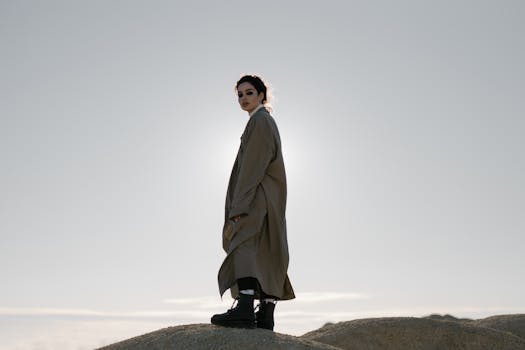 A woman stands on a dune in a trench coat, fashionably silhouetted against the sunlight.