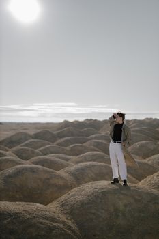 一名女子穿着时尚风衣，站在岩石沙漠地形上，明亮的阳光照耀着她