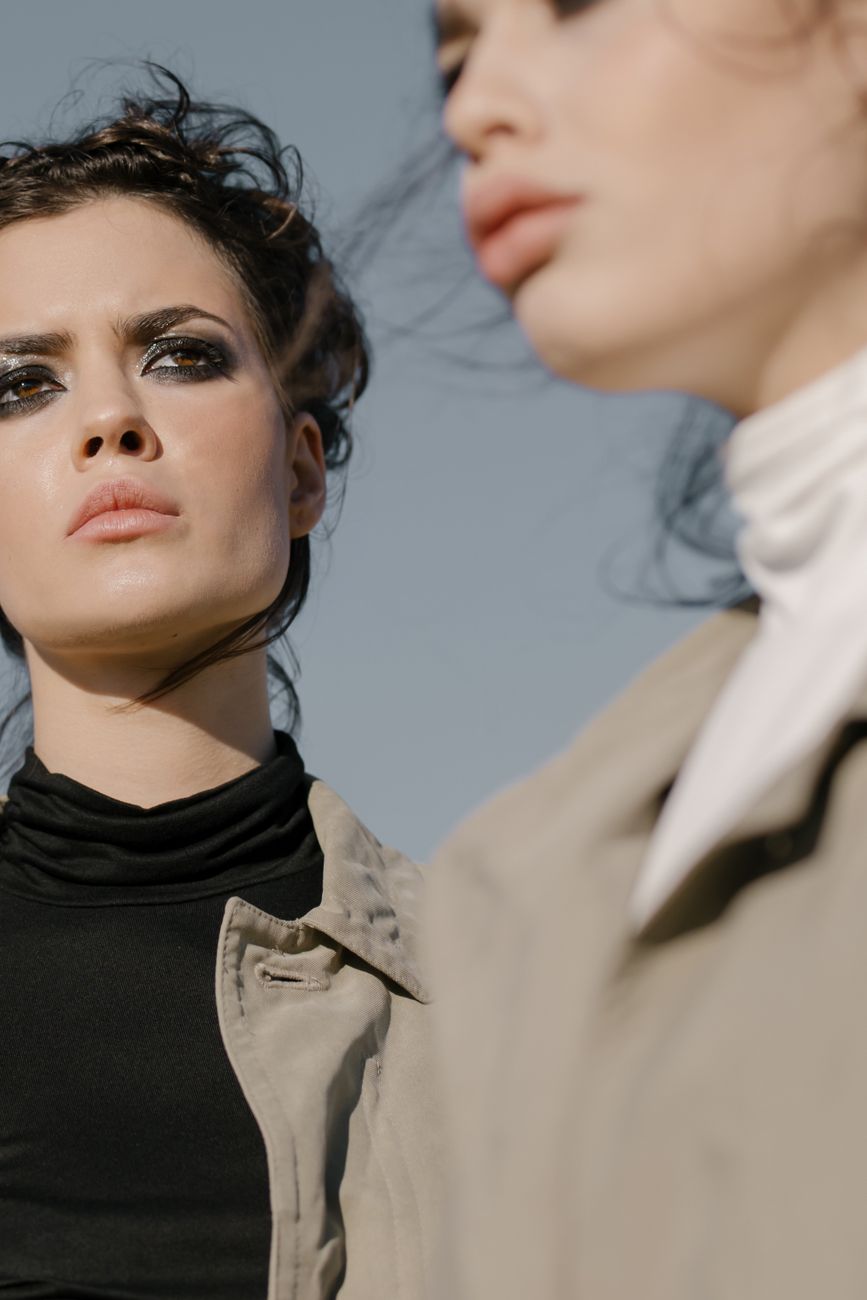 Close-up portrait of two stylish women with bold makeup and fashionable attire, shot outdoors.