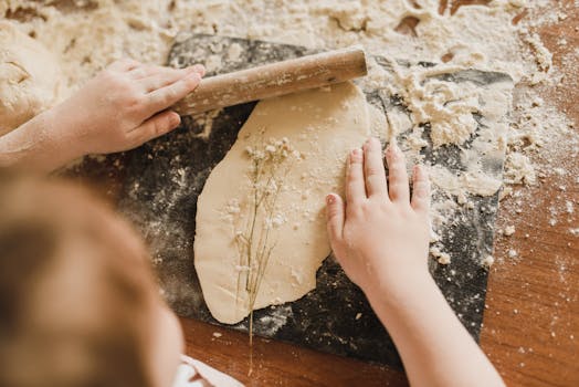 Close-up of a child rolling dough with hands and rolling pin on a floured surface indoors.
