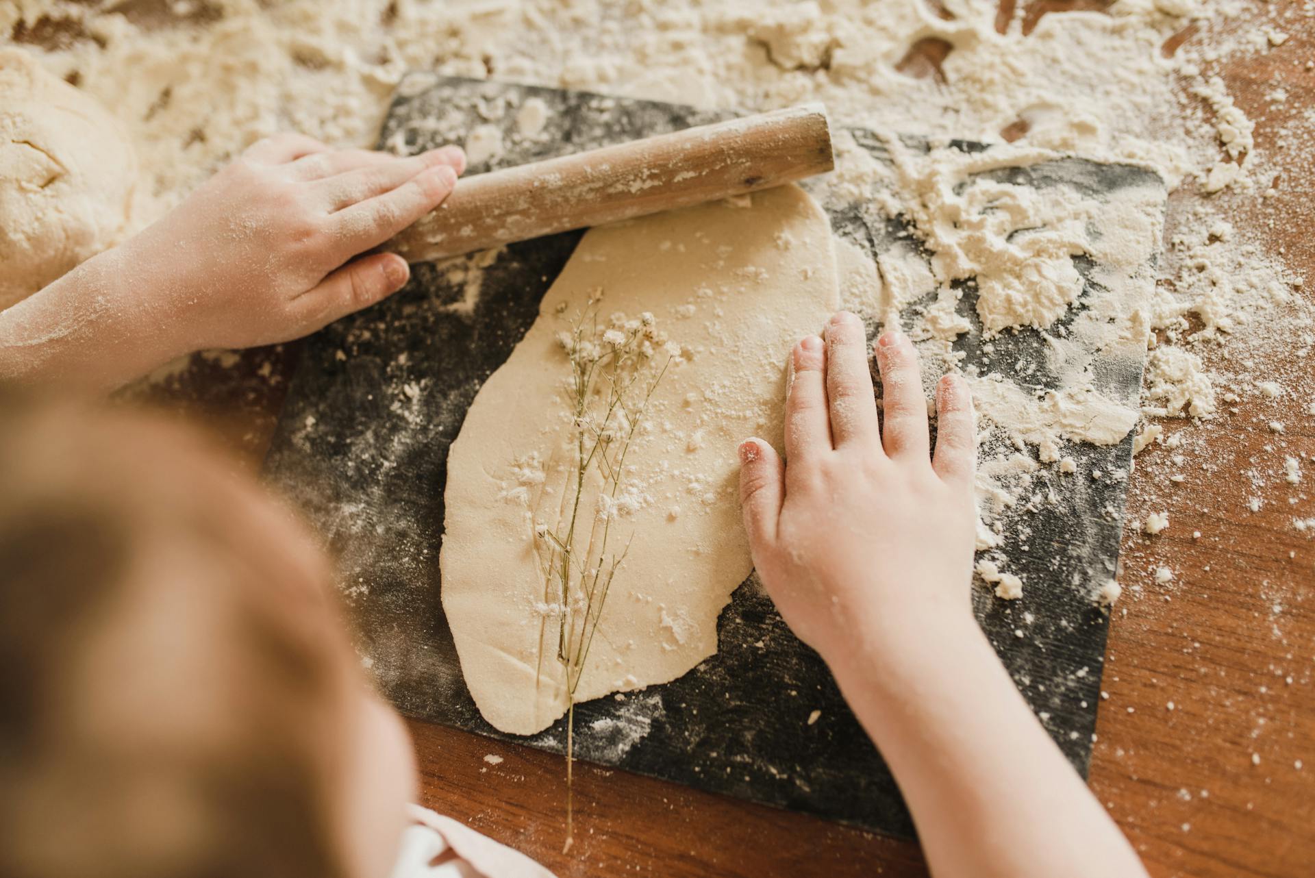 Close-up of a child rolling dough with hands and rolling pin on a floured surface indoors.