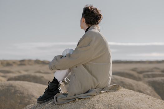 一位穿着风衣和黑色靴子的女人坐在山上，凝视着沙漠的地平线