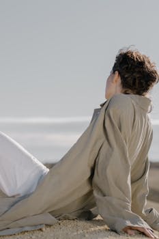 一名女子穿着米色外套和白色裤子，坐在沙地上，在户外享受宁静的一天