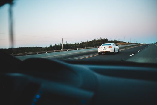 Through windshield of car with black car interior driving on asphalt roadway along green trees in daylight against cloudless sky