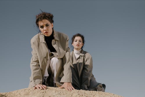 Two Women Wearing Trench Coats Sitting on Sand