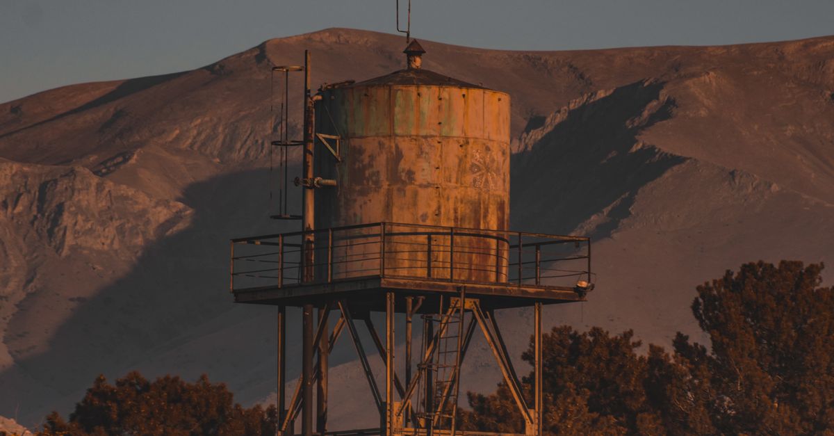 Photo by Ahmet Polat A weathered metal water tank stands tall amidst green trees with a mountain backdrop.