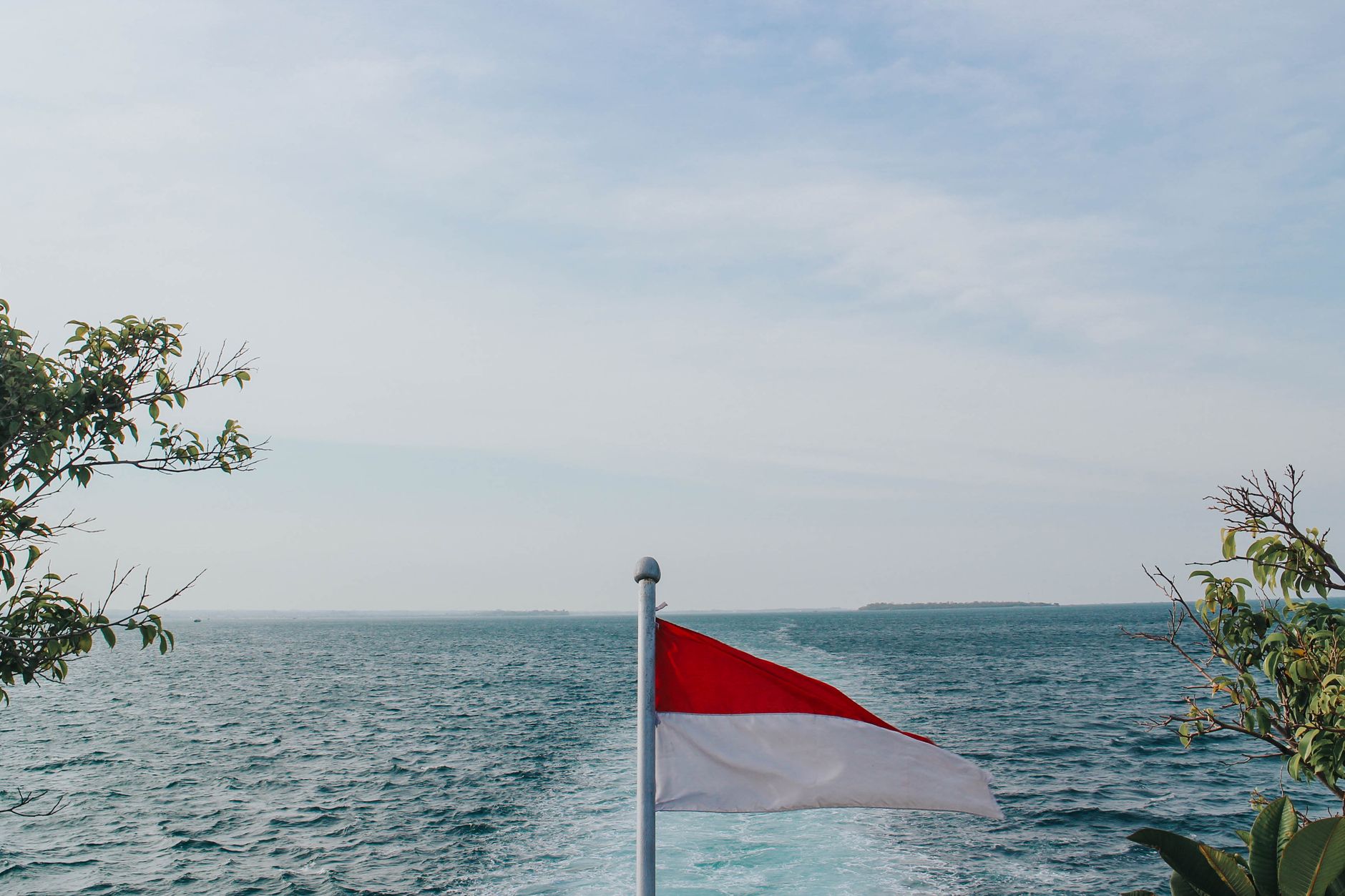 A red and white Indonesian flag above a vast ocean horizon with clear sky.
