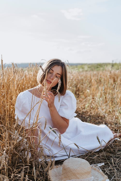 Woman in White Dress Sitting on Wheat Field Under Blue Sky