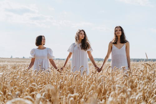 Three Women in White Dresses Holding Hands in a Wheat Field