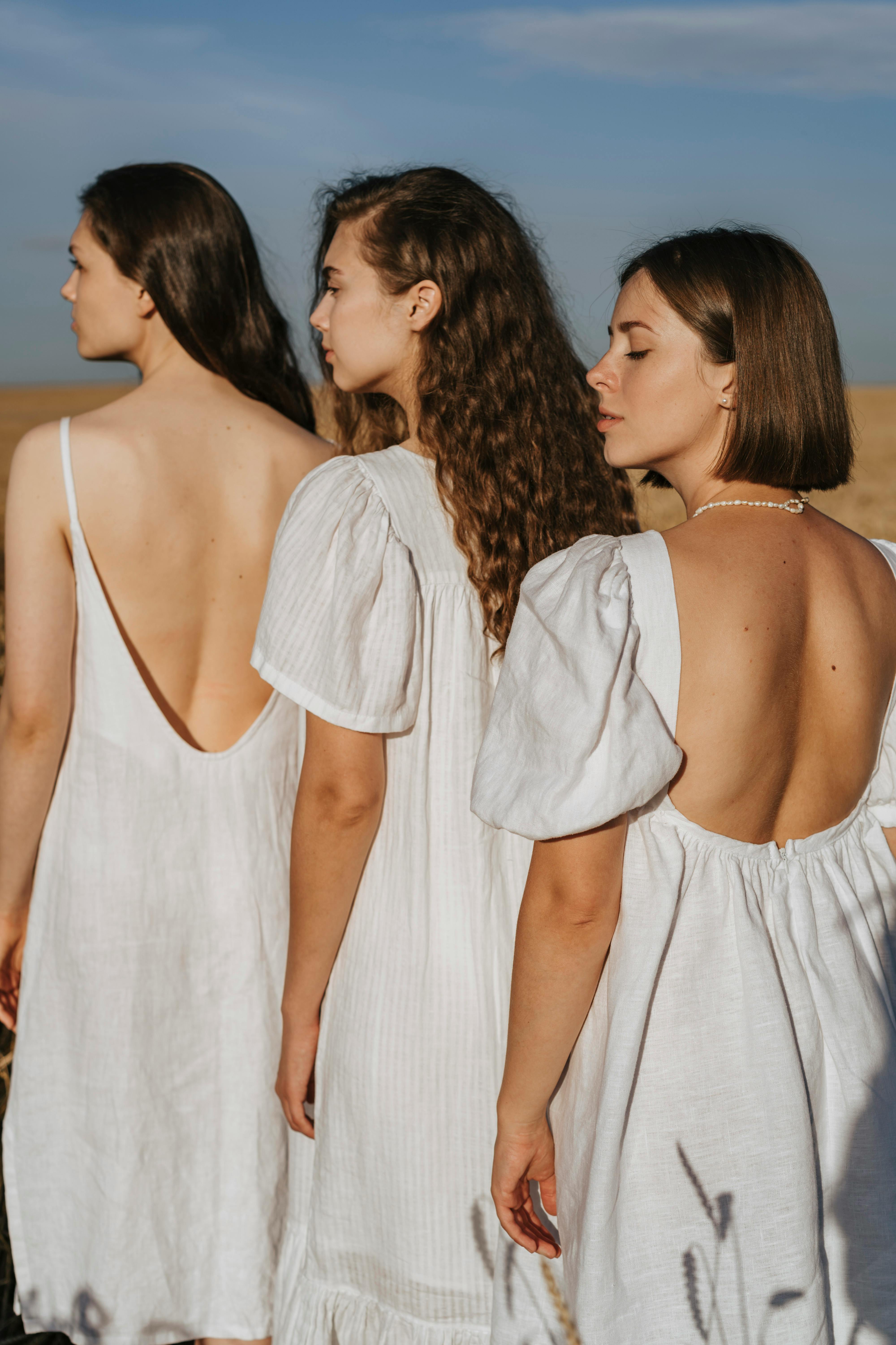 Three women in white dresses standing outdoors with eyes closed, embracing nature's serenity.