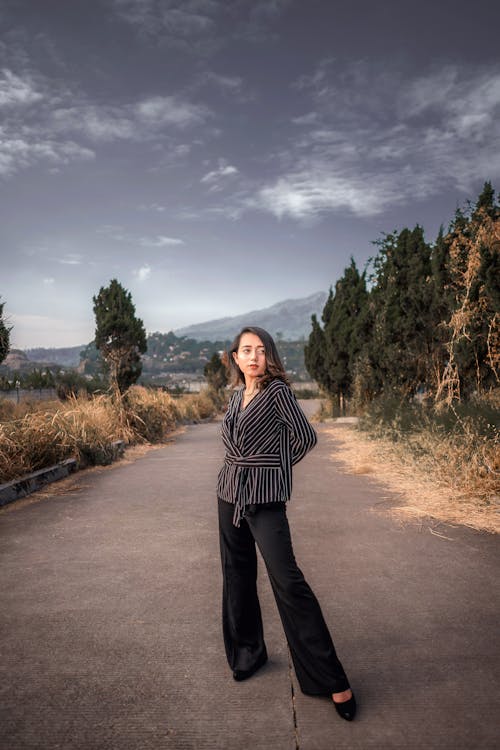 Free Full body of confident young female in stylish suit standing on asphalt road near forest with hand behind back against cloudy sky Stock Photo