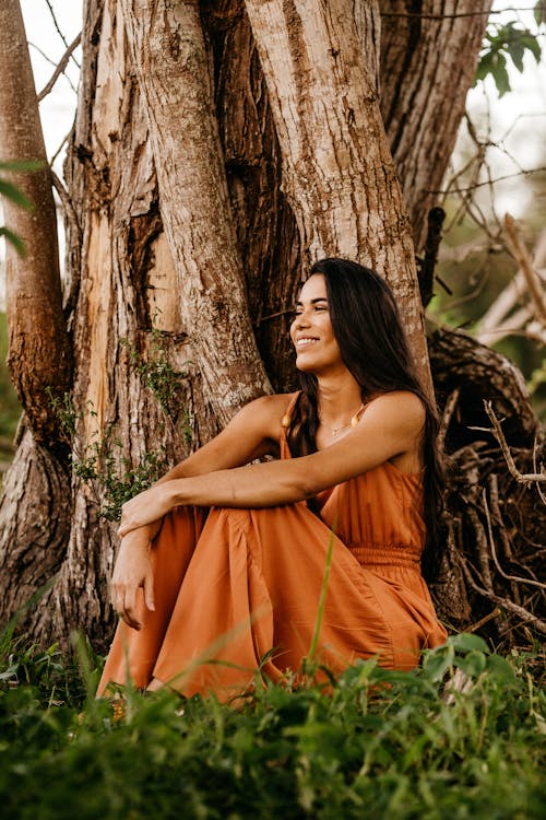 Full body cheerful young brunette in maxi summer dress relaxing on grassy lawn and leaning on tree while embracing knees and looking away with radiant smile