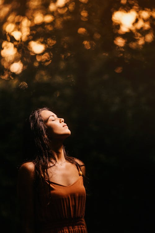 Tranquil attractive brunette in brown dress standing with eyes closed against blurred green nature background and turning face to light