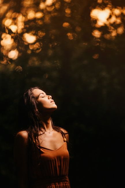 Tranquil attractive brunette in brown dress standing with eyes closed against blurred green nature background and turning face to light