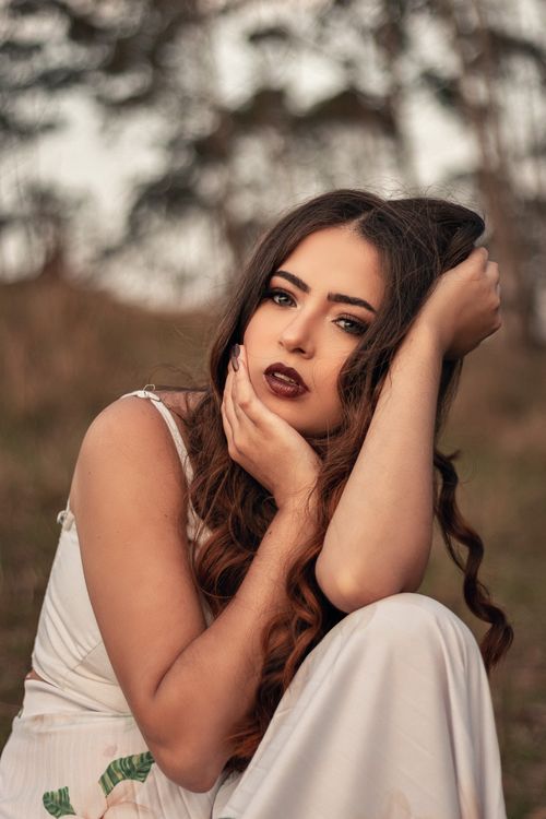 Free Calm female with dark hair wearing white dress looking at camera and sitting in nature with blurred trees while tousling hair and hand on face Stock Photo
