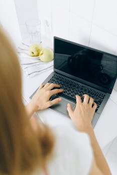 Overhead view of hands typing on a laptop with apples and glass on a white desk.