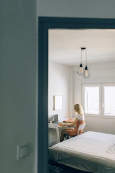 A woman works on her laptop in a bright minimalist bedroom, viewed from the doorway.