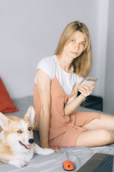 一位年轻女子坐在床上，拿着智能手机，和她的柯基狗一起在室内享受休闲时光