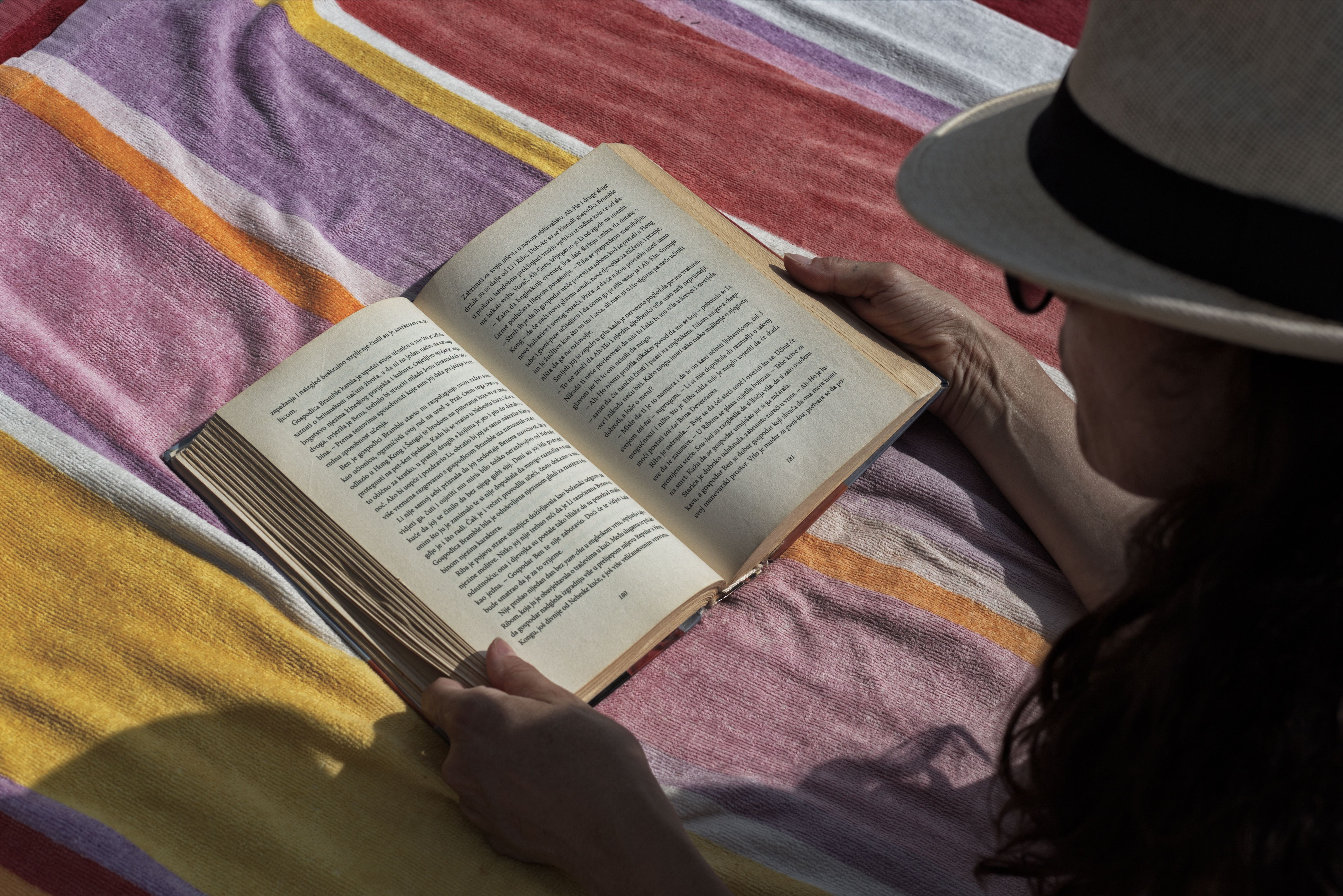 High angle of crop female in straw hat enjoying book while lounging on plaid in sunlight