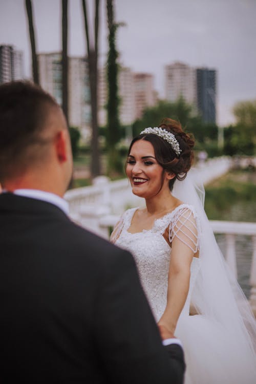 Cheerful bride in elegant wedding dress with hairdo and makeup smiling happily while holding hand of groom