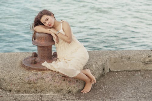 From above full body of barefoot resting on waterfront while leaning on bollard and looking at camera