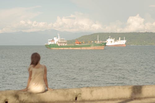 Free Back view of unrecognizable female sitting near rippling sea with floating ship in sunny summer day Stock Photo