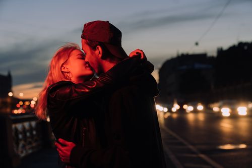 Free A couple sharing an intimate kiss on a city street during twilight, with blurred lights in the background. Stock Photo