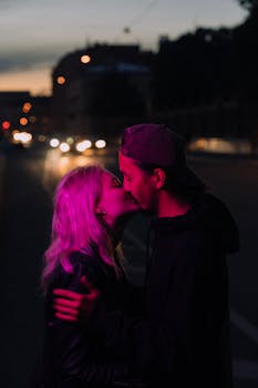 A young couple shares a tender kiss on a city sidewalk at night, illuminated by streetlights.