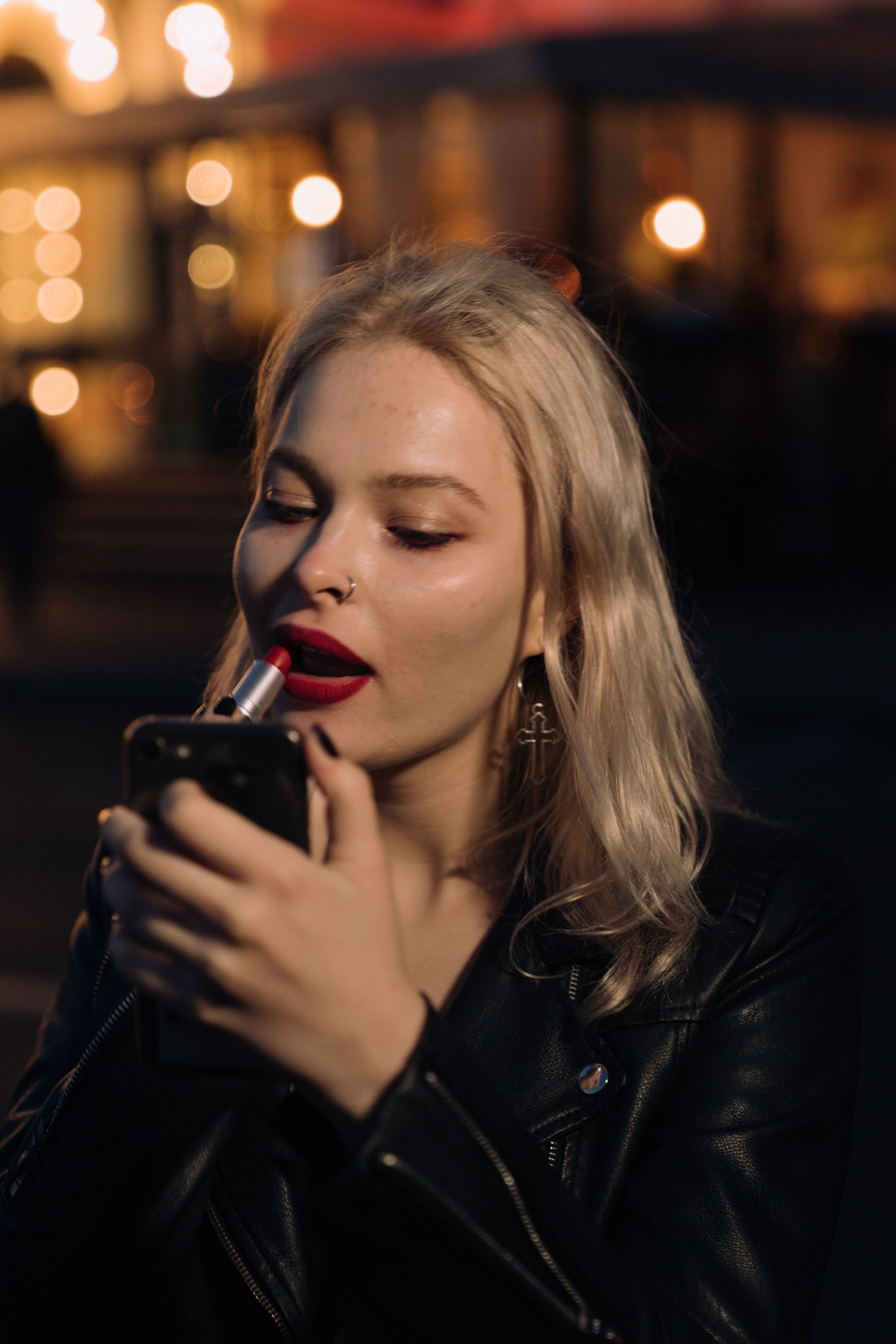 晚上，一位穿着皮夹克的金发女子用智能手机当镜子涂口红