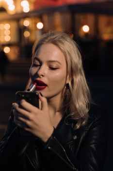 晚上，一位穿着皮夹克的金发女子用智能手机当镜子涂口红