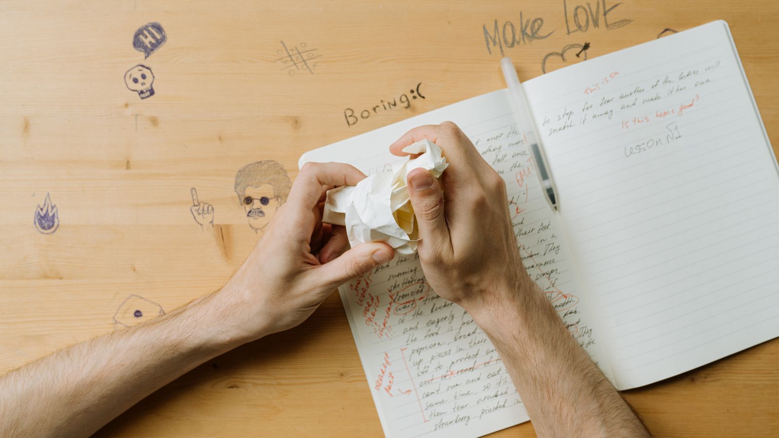 Hands crumpling a sheet of paper above an open spiral notebook on a wooden table