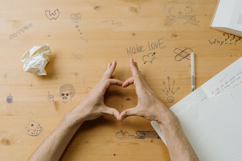 Free Hands form a heart shape on a doodle-covered wooden desk, adding a playful touch. Stock Photo