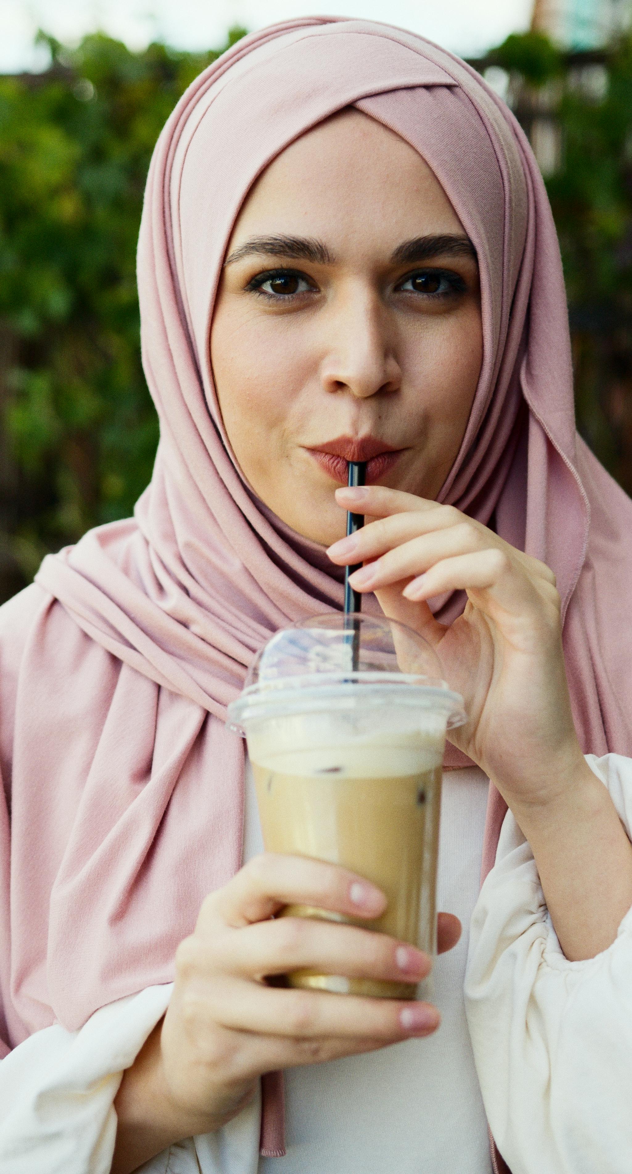 woman in pink hijab holding clear plastic cup with brown liquid