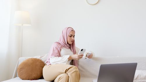 Free Young woman in hijab sitting on a couch using a smartphone with a laptop nearby. Stock Photo