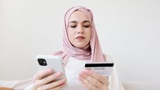 一名女子戴着头巾，坐在室内用智能手机和信用卡进行网上购物