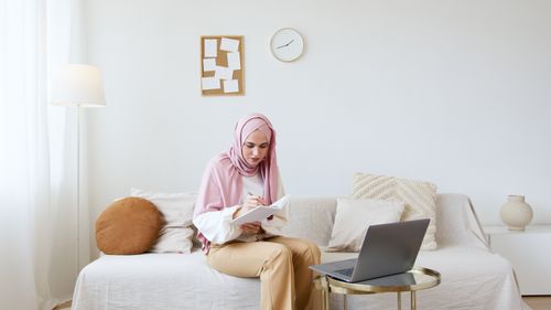 Free Woman in hijab sitting on sofa, writing in notebook with laptop nearby. Stock Photo