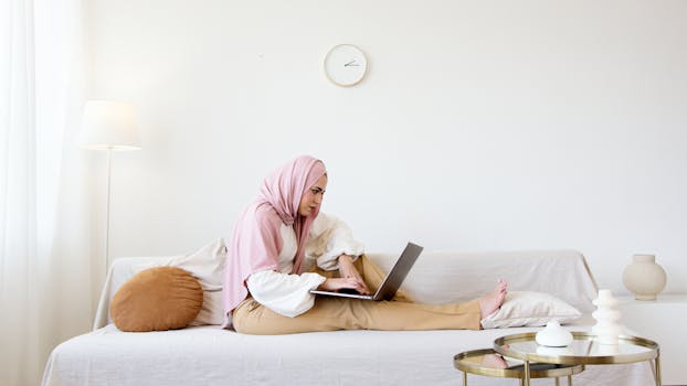 A woman in a hijab relaxes on a sofa while working on her laptop in a minimalistic living room.