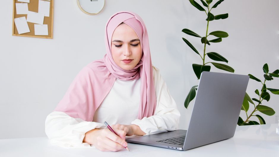 A woman in a hijab writing at a desk with a laptop and indoor plant.