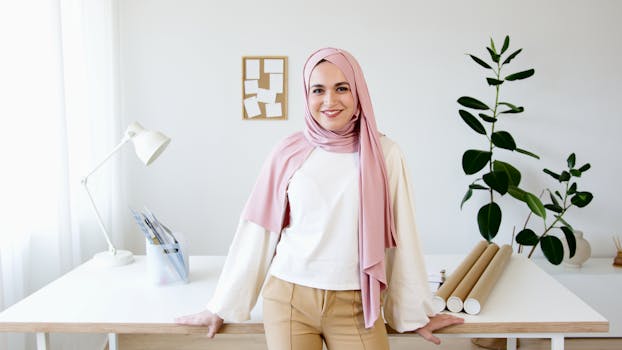 A smiling woman in a pink hijab stands confidently by a desk in a modern well-lit office setting.