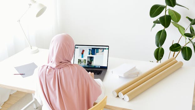 A woman wearing a hijab is working on her laptop at a modern desk with indoor plants and natural light.