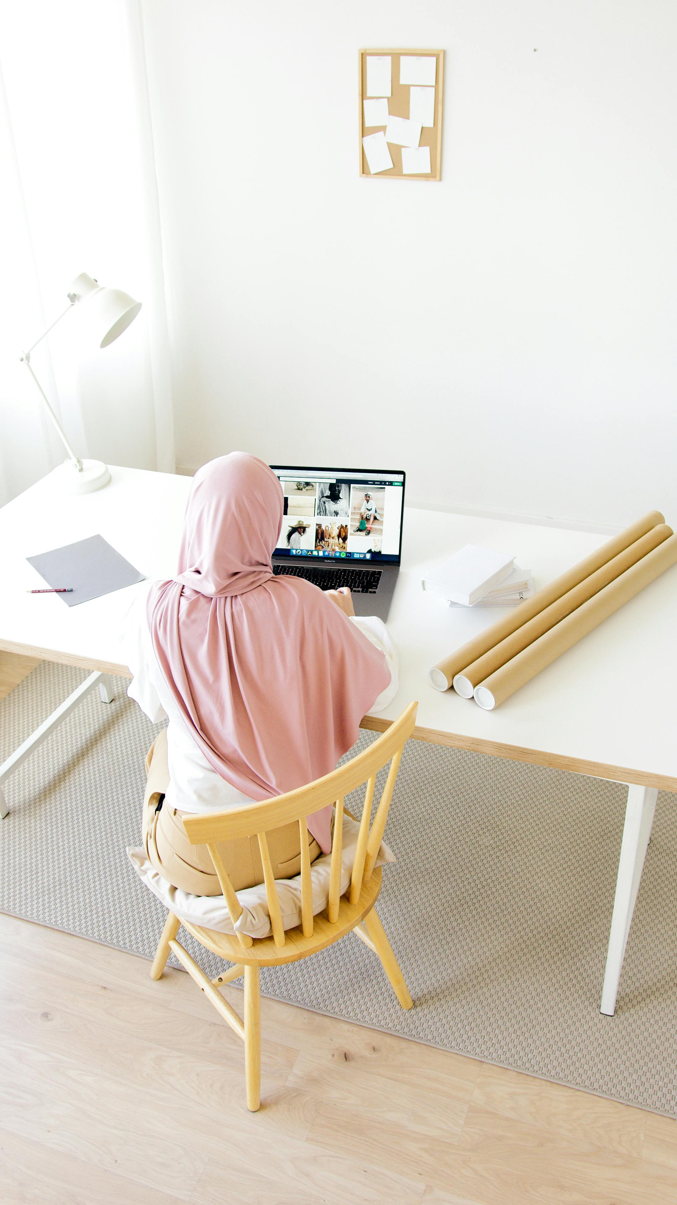 a woman in pink hijab sitting on a wooden chair while using her laptop
